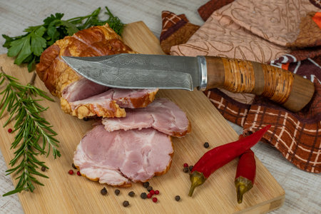 On a wooden cutting board, sliced juicy pork ham, carving knife, red hot peppers, multi-colored allspice, parsley, dill and basil. The concept of delicious pork productsの写真素材