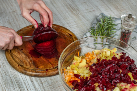 Red boiled beets are cut into small pieces with a kitchen knife on a wooden cutting board, next to it are a bowl of meat salad being prepared, a pepper grinder, a sprig of rosemaryの写真素材