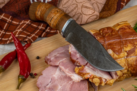 Sliced slices of juicy pork ham on wooden cutting board, next to it are a carving knife, multi-colored allspice peas, red hot peppers, parsley, dill and basil. The concept of delicious meat products.の写真素材
