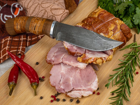 On a wooden cutting board, sliced juicy pork ham, carving knife, red hot peppers, multi-colored allspice, parsley, dill and basil. The concept of delicious pork productsの写真素材