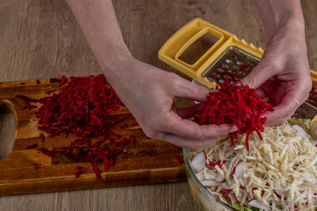 Close-up of female hands grate red beets on an iron grater for salad preparation on a wooden cutting board. Preparing fresh vegan vegetable salad. The concept of healthy eating as a lifestyleの写真素材