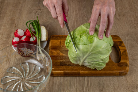The female cuts cabbage on a wooden cutting board for cooking vegetable salad from fresh raw vegetables cabbage, radish, green onion, red beet, celery root. Vegetarianism and a healthy lifestyleの写真素材