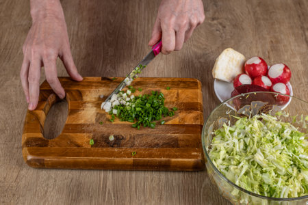Close-up of womans hands chopping green onions with a kitchen knife for making salad on a wooden cutting board. Preparing fresh vegan vegetable salad. The concept of healthy eating as a lifestyleの写真素材