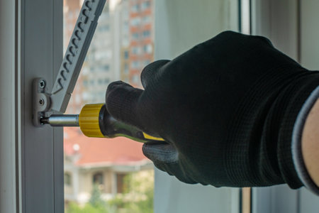 The worker with a screwdriver screws the fixing screw into the window frame, setting the window opening regulator. Installation of a limiter on a plastic window frame. Window guard fixtureの写真素材