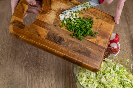 The cook puts chopped green onions from a wooden cutting board into a bowl of vegetable salad. Close-up of cooking fresh vegetable salad in home kitchen. The concept of natural food useful to healthの写真素材