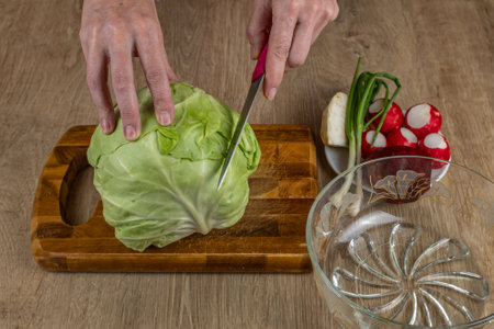 A woman uses a kitchen knife to cut fresh white cabbage on a wooden cutting board. Close-up female hands chopping cabbage for making summer salad. The concept of cooking food in the home kitchenの写真素材