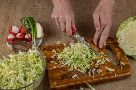 A woman uses a kitchen knife to cut fresh white cabbage on a wooden cutting board. Close-up female hands chopping cabbage for making summer salad. The concept of cooking food in the home kitchenの写真素材