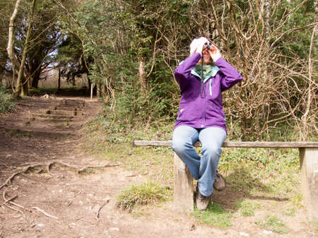 Woman sitting on a wooden bench in the countryside looking through a pair of Binocularsの写真素材
