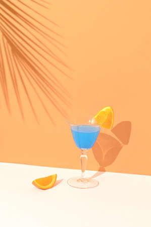 Blue cocktail drink decorated with a slice of orange. In the background shade of tropical palm leaves on a combination of light brown and cream background. Summer vibes party concept.の写真素材
