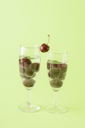 Two glasses of champagne filled with a cherry drink on a green background. One cherry between two glasses as a connection.の写真素材
