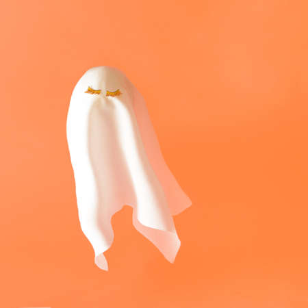 White ghost costume with gold lashes flying on a orange background. Halloween minimal composition.の写真素材