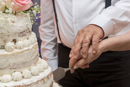 Older couple dressed luxuriously cutting together a beautiful three tier wedding anniversary cake. Concept of union and eternal love. Selective focus.の写真素材
