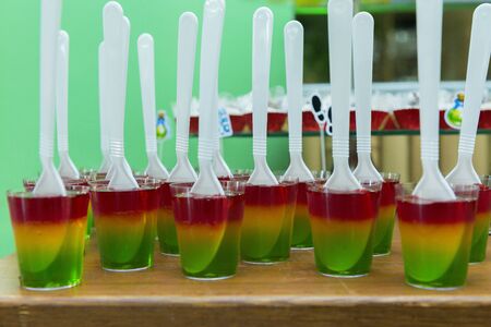 Glass with colorful striped jelly above wooden table. Rainbow jelly for kids party. Plastic spoon in...