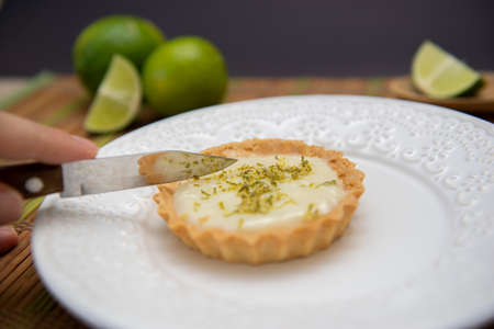Close up of knife cutting lemon tart on dish plate with lemon slices. Lemon tartlet with zest on...