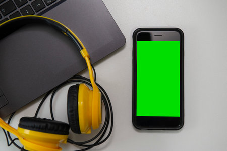 Top view of office desk with open laptop, headphones and smartphone with green screen. Close up to insert image on cell phone screen. Selective focus on the cell phone.のeditorial素材