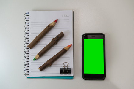 Top view of office desk with blank note book, rustic colored pencils and smartphone with green screen. Close up to insert image on cell phone screen. Selective focus on the cell phone.のeditorial素材