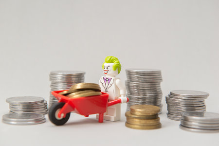 Florianopolis, Brazil, March 28, 2020: Lego Joker minifigure stealing money, carrying coins on a wheelbarrow on white background. Selective focus. Lego minifigures are manufactured by The Lego Group.のeditorial素材