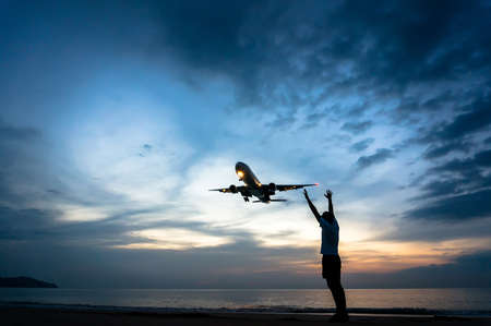 a man stands on the beach and stretches his hands to the airplane flying above him, in the evening...