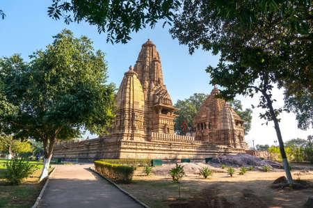 Old Hindu temple, built by Chandela Rajputs, at Western site in India's Khajuraho framed by trees. White grey for the younger and older beige structure against blue skies over green grass.の写真素材