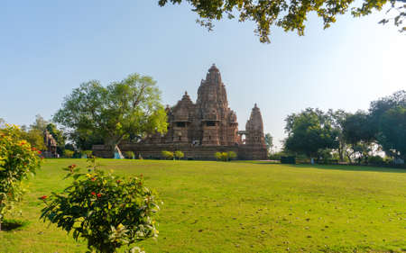 Old and younger Hindu temple, built by Chandela Rajputs, at Western site in India's Khajuraho framed by trees. White grey for the younger and older beige structure against blue skies over green grass.の写真素材