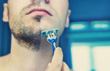 Young man shaves his short beard shaving machine tool. Half face with a beard half shaved.の写真素材