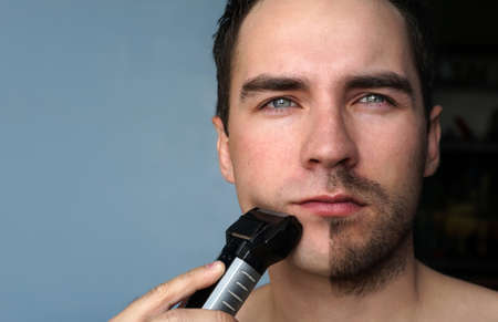 young man shaving his beard with electric shaver. He shaved half of beard on grey blue background