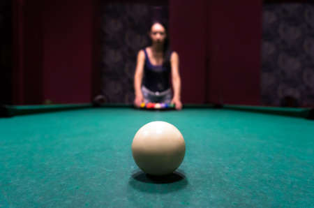 Young brunette girl puts balls on the billiard table. Big white billiard ball in the foreground.
