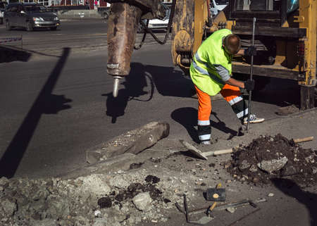 Worker repairing a road hammer crowbar asphalt next to the excavator on the background of a city streetの写真素材