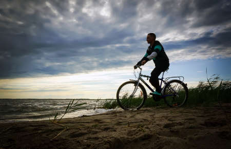 Image of sporty one tourist man walking along the shore with the bike against sunset sky with clouds and sun rays.の写真素材