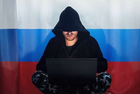 Hacker in a dark hoody sitting in front of a notebook on the background of the Russian flag. Russian hacker intervenes in the American and European elections.の写真素材