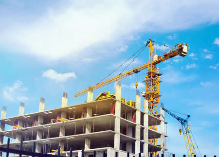 High yellow crane building a house in the beautiful bright blue sky with clouds. Workers in the building under construction is building a house.の写真素材