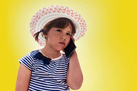 Little brooding girl in a pink hat and a striped dress talking on smartphone . little fashionista isolated on yellow backgroundの写真素材