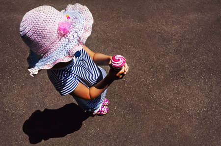 Top view of little girl in pink hat eating ice cream on the empty gray background of asphalt. Little fashionista in trendy clothes in the summerの写真素材