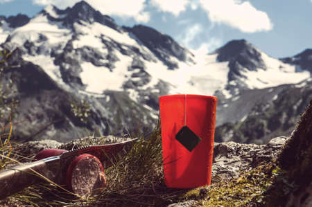 Orange plastic mug with strong tea and tea bag on a snowy winter background. Tourist knife cuts smoked sausage.の写真素材