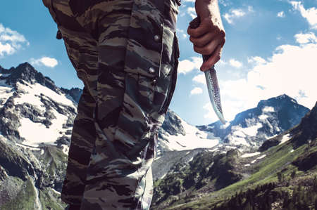 Combat knife in hand the soldier the commando in the background of snow-capped peaks in the mountains. Scout with a knife. Tourist backpacker holds the knife in the wild.の写真素材
