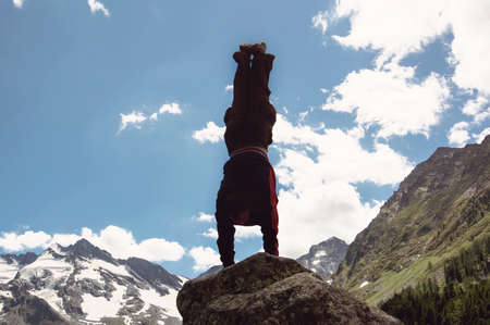 man doing complex Yoga exercise headstand. Amazing Yoga landscape in beautiful mountains. Dangerous stunts traceur standing on his hands on the edge of a cliff.の写真素材