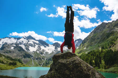Athletes gymnast do dangerous exercises on rock on background of beautiful mountain scenery with snow-capped peaks and turquoise lake and blue sky with white clouds. Practice gymnastic exercises in the open air in natureの写真素材