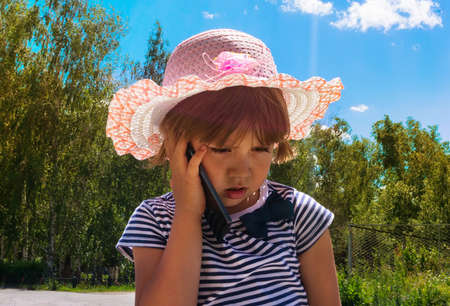 Kid with smartphone with a thoughtful face. little baby girl cell phone talking to her mom, dad family on green trees outdoors summer background. Kids and technology conceptの写真素材