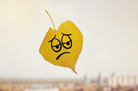 Yellow leaf with a picture of a sad face on the glass window in the blurred background city. The autumn has come. Fall outside the window.の写真素材
