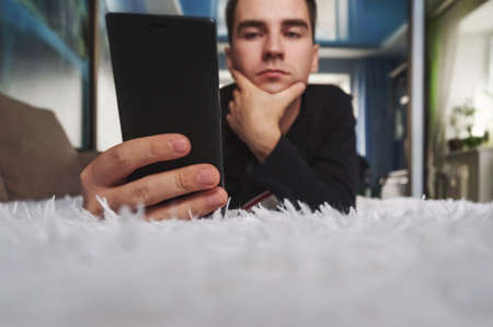 a young man looks at his smartphone at home. Man relaxing during looking at the screen while lying on sofa. Read electronic books on your phone.の写真素材