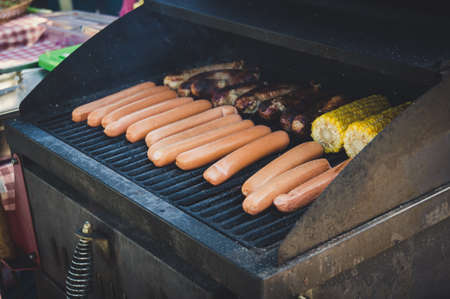 Fresh sausage. Hot dogs grilling outdoors on a gas barbecue grill with vegetables eggplant with corn and potatoesの写真素材