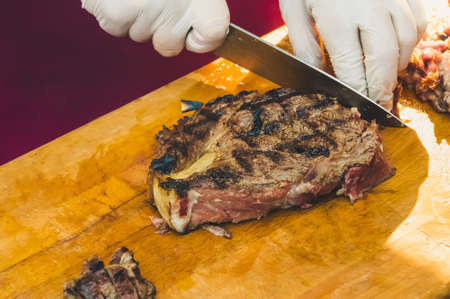 Male chef cutting big piece of beef on wooden board in restaurant kitchen. Man cook preparing steak and vegetable garnish. Hands cooks in white clean sterile gloves close-up.の写真素材