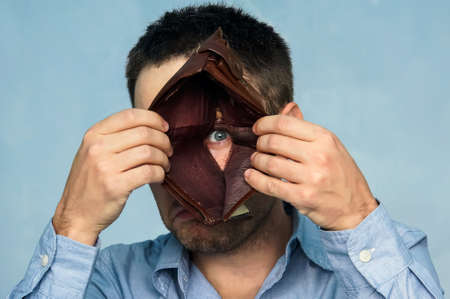 Closeup portrait, stressed, upset, sad, unhappy young man standing with, looking into hole of empty wallet. Financial difficulties, bad economy concept. Negative emotionの写真素材