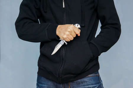 A drug addict burglar in a black hoodie. The concept of the crime of robbery. Closeup of a young man hand, holding a knife, about to attack, mafia.の写真素材