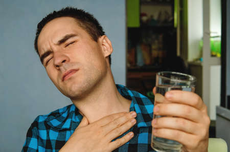 Pain and sore throat. Handsome guy in casual clothes is holding a glass of water and touching his throat while sitting on couch at home. A parched throat and a glass of water in handの写真素材