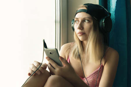 Pretty blond woman listening music, at home, having fun. beautiful woman listening music in black headphones and sitting on windowsill. Female wear pink sexy clothesの写真素材