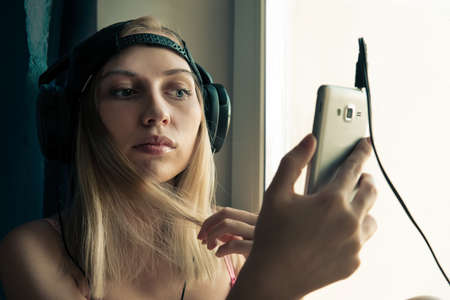 Young blonde woman listening to music on headphones while sitting by window. The girl stares at the...