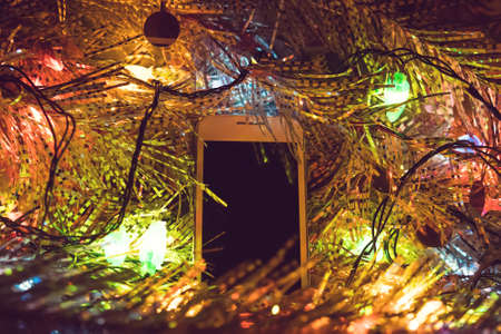 smartphone with gifts and decorations in front of Christmas tree. Focus on smartphone. White phone in glowing Christmas lights. Blank screen for photo or text. copy spaceの写真素材