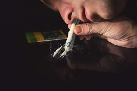 Young addict snorting white cocaine through a hundred dollar bill on black background. concept of addictionの写真素材