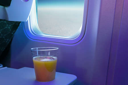 A cup of orange juice on tray at cabin seat of an aircraft. Glass of drink on the background of the window of the aircraft. Flying above the clouds.の写真素材